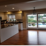 An open-concept white kitchen and dining area with reddish-brown wood floors and a view of a pool through glass sliding doors.