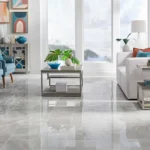A bright, professionally styled living room with high-gloss gray marble-look floor tiles and blue accent chairs.
