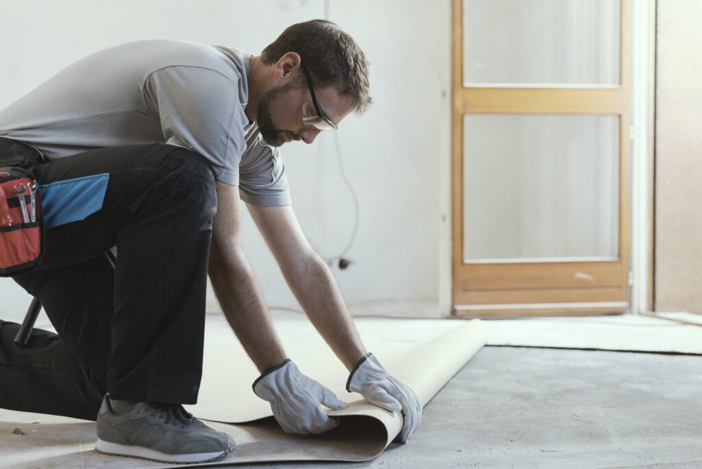 A worker wearing safety glasses and gloves kneeling on a concrete floor to roll out a tan underlayment or linoleum sheet.