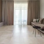 A modern living room with large, matte-finish cream floor tiles and floor-to-ceiling beige curtains.