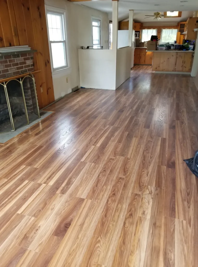 An open-concept living area with newly installed light-brown wood laminate flooring, a brick fireplace, and wood-paneled walls.